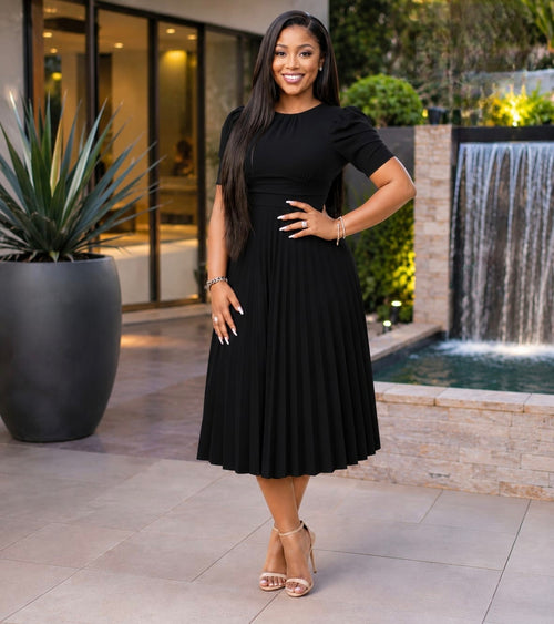 Black Pleated Elegance Midi Dress with structured bodice and puffed short sleeves, styled with strappy heels near greenery.