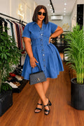 Model wearing a denim button-up flare mini dress with short sleeves and a collared neckline, accessorized with sunglasses and a handbag.