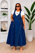 Model wearing a dark denim wrap overall maxi dress with wide adjustable straps and a large patch pocket over a fitted white tee.