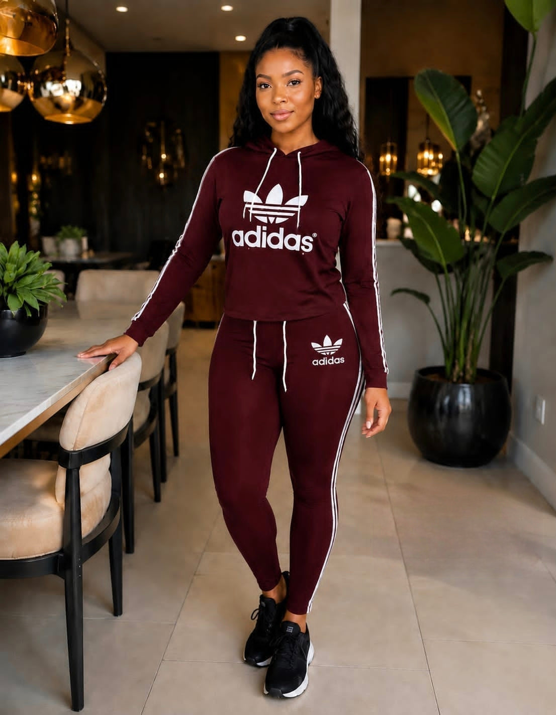 Burgundy Adidas tracksuit set featuring a hoodie with drawstring closure and joggers with side stripes, modeled indoors.